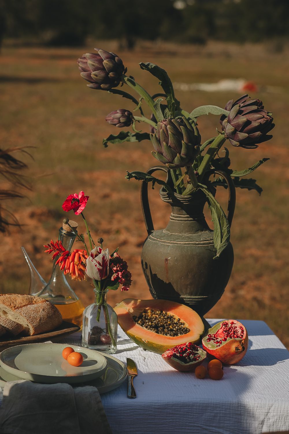 Rustic outdoor table setting with artichokes, pomegranate, and flowers in earthen vase.
