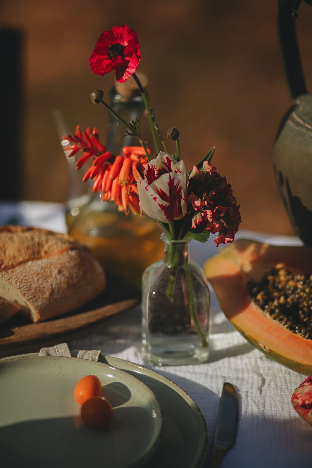 Red carnations and wildflowers in glass vase on table with fruit and bread.