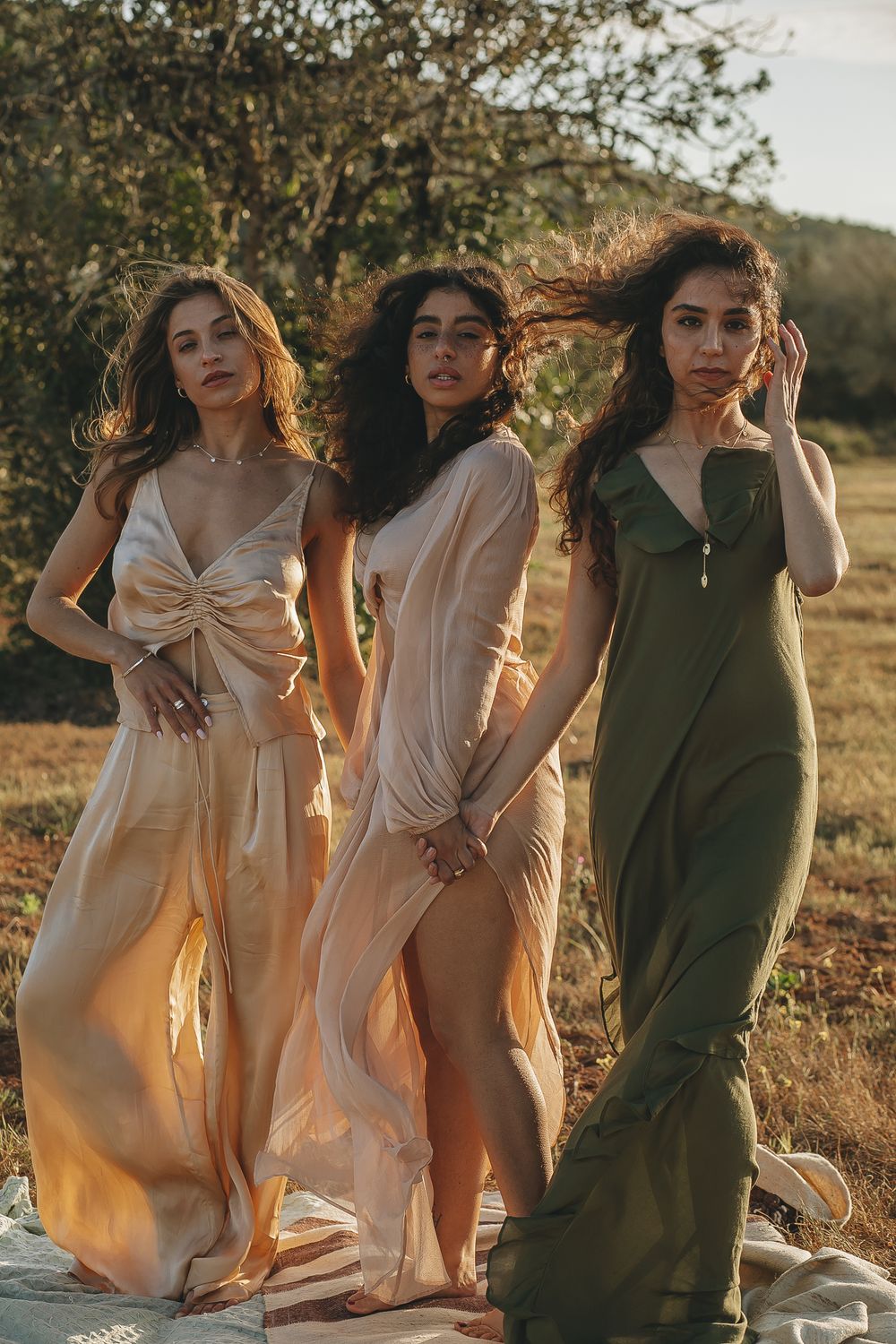 Friends in light colored dresses posing together in a grassy field during sunset.