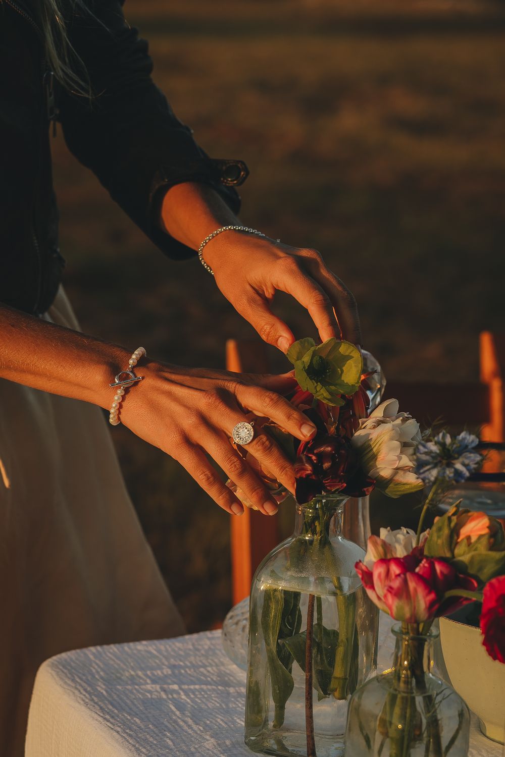Hands arranging colorful flowers in glass vases on a white tablecloth at sunset.