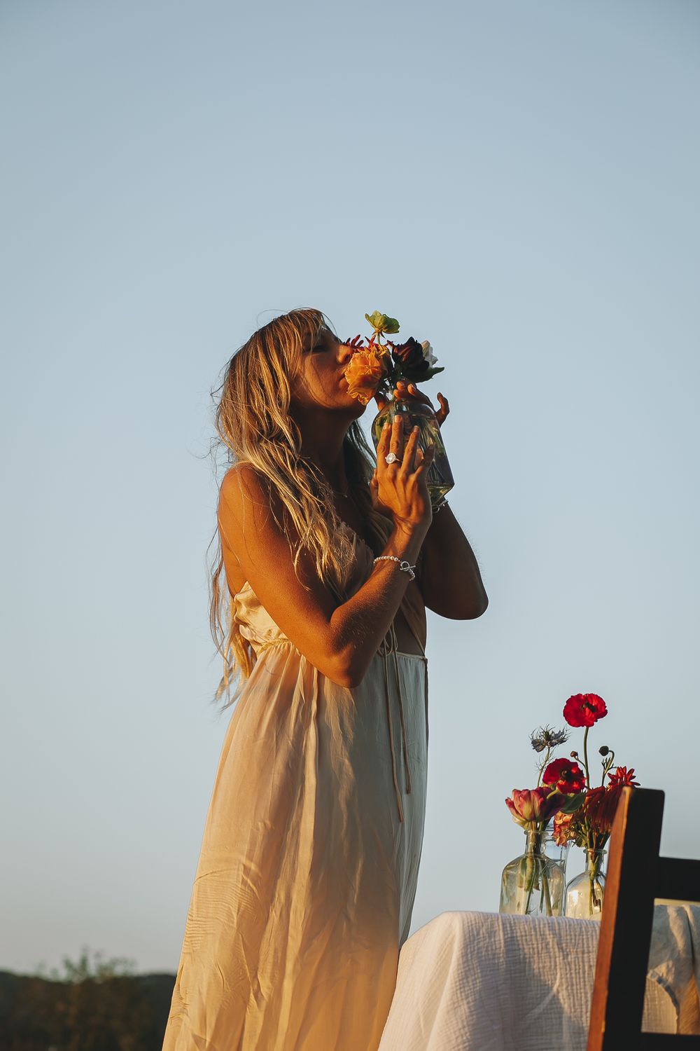 Figure in flowing white dress photographing flowers against a pale blue sky.