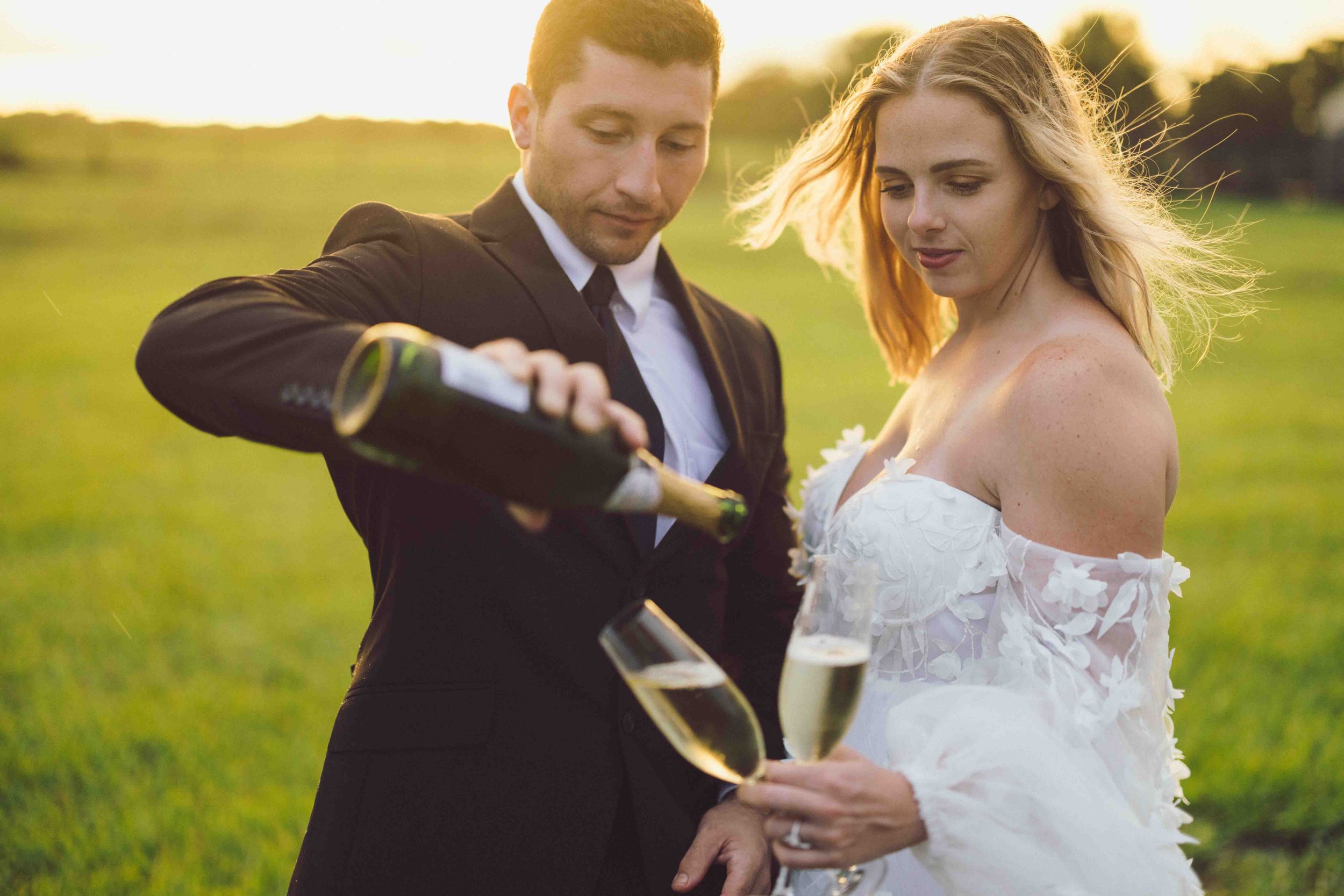 A couple opens champagne together in a sunlit field during their wedding celebration.