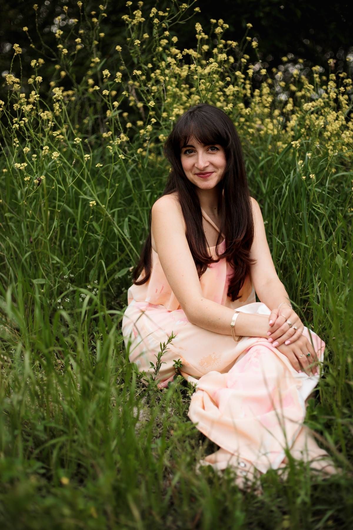 A woman with long dark hair and a peach dress sits in tall green grass beside yellow wildflowers, smiling softly.