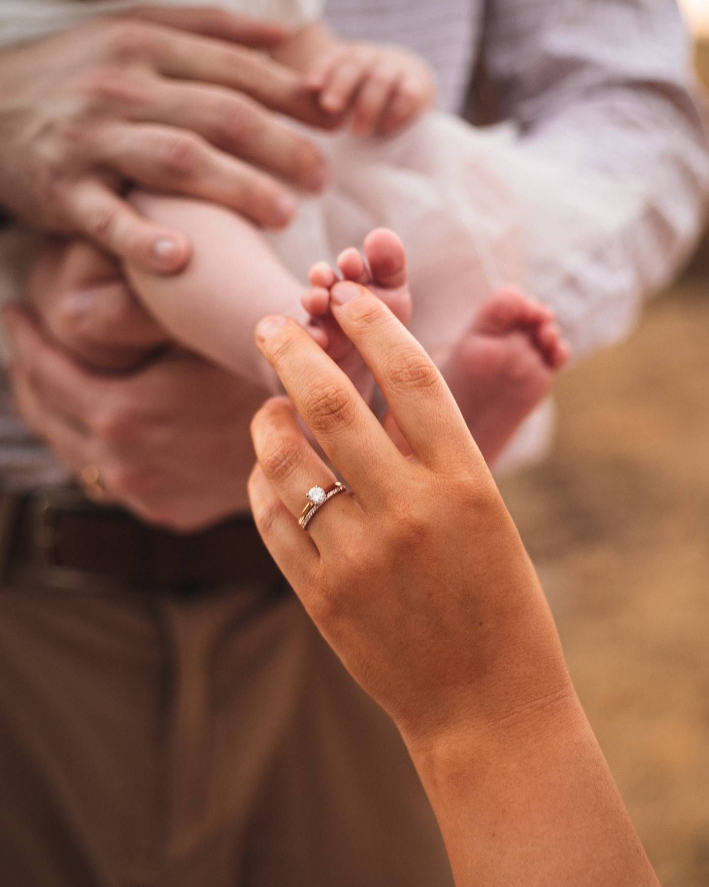Woman's hand with a diamond engagement ring reaches toward a baby's tiny feet held by a parent in a cozy family photo.