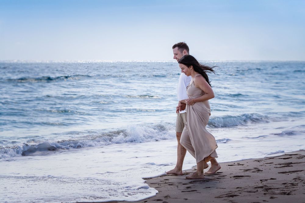 Romantic beach photoshoot with couple walking along shoreline in white outfits at sunset.