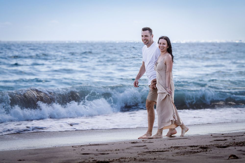 Couple enjoying a romantic stroll on sandy beach with crashing waves in background.
