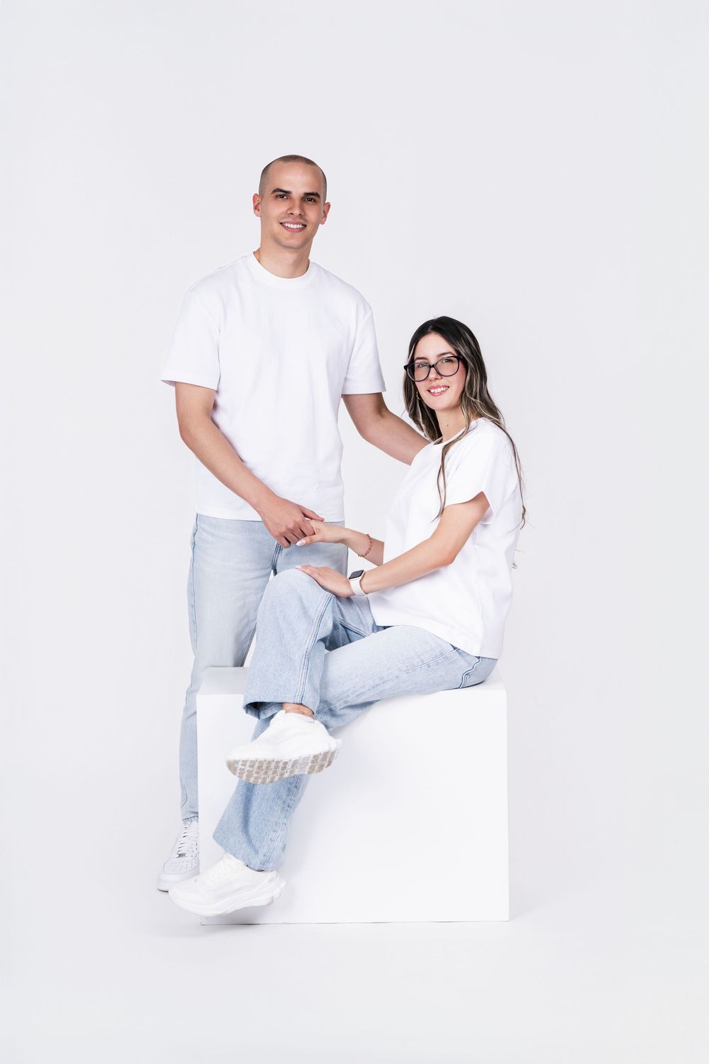 Modern couple in matching white shirts and jeans posing on white cube against plain background.