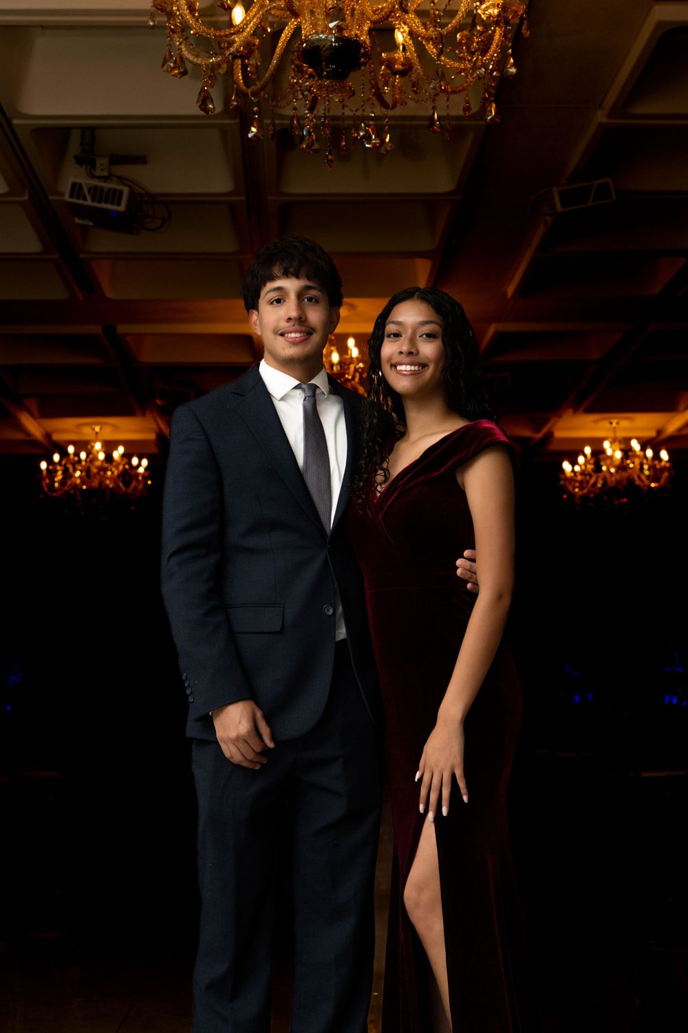 Elegant couple poses under ornate chandeliers in formal evening attire.