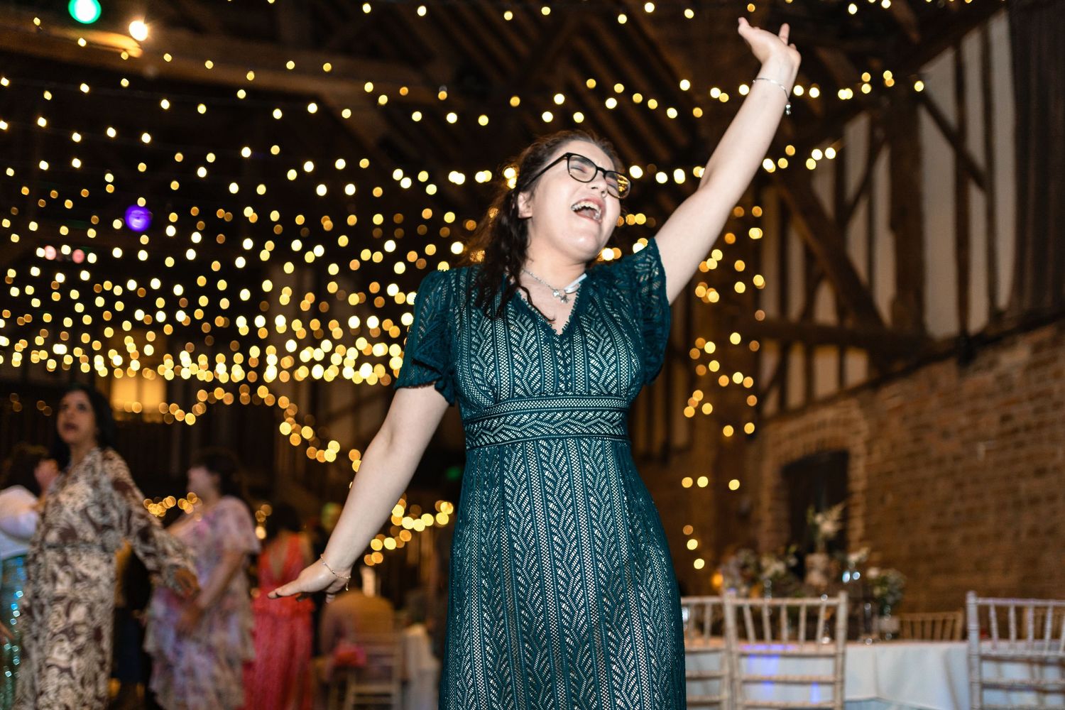 People dancing and celebrating at an indoor wedding reception venue decorated with twinkling string lights and brick walls.