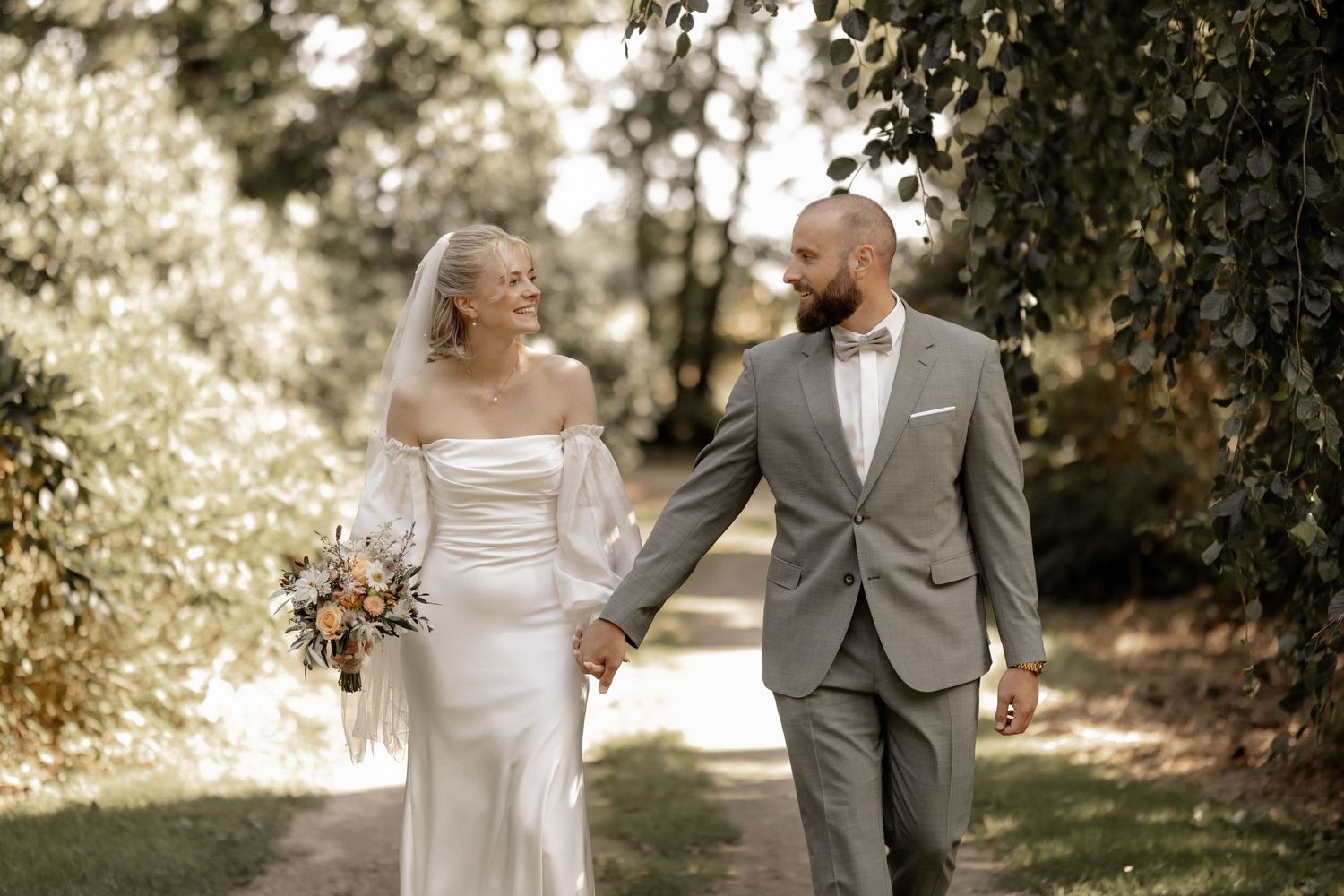 Bride and groom walking hand in hand along a sunlit garden path, bride holding a colorful bouquet.