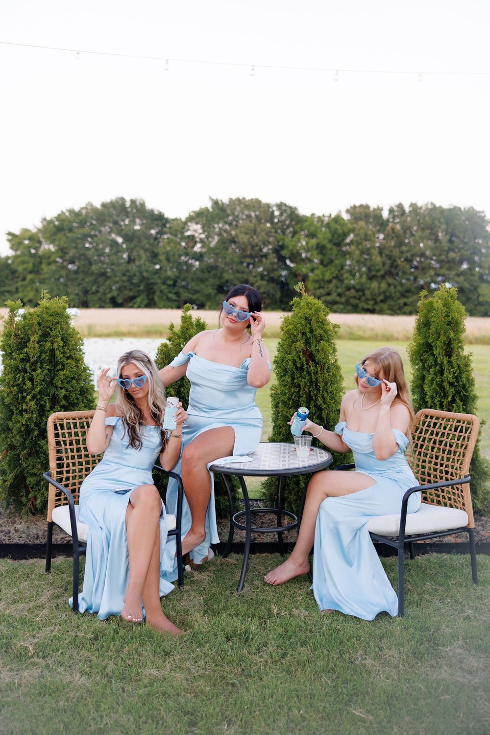 Three women in light blue satin gowns sit outdoors at a small table, wearing fun sunglasses and enjoying a celebration.