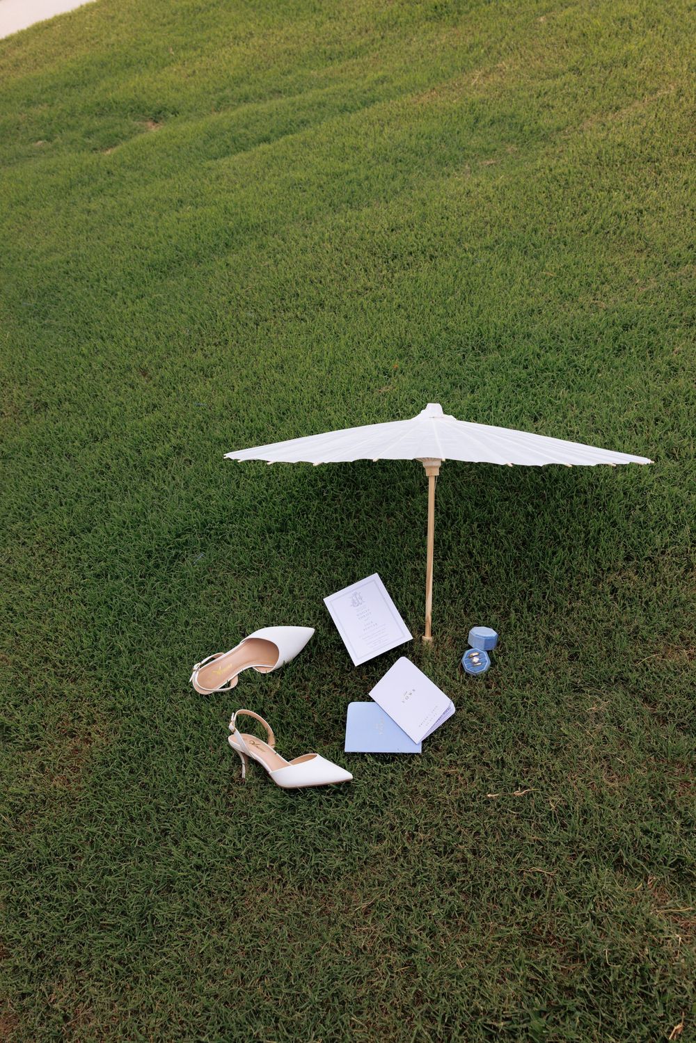 Wedding accessories arranged on green grass under a white parasol, including heels, rings, and stationery.