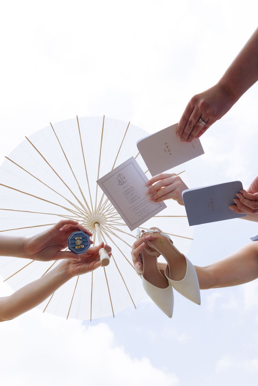 Hands holding a white parasol, wedding program, and accessories against a bright sky at an outdoor ceremony.