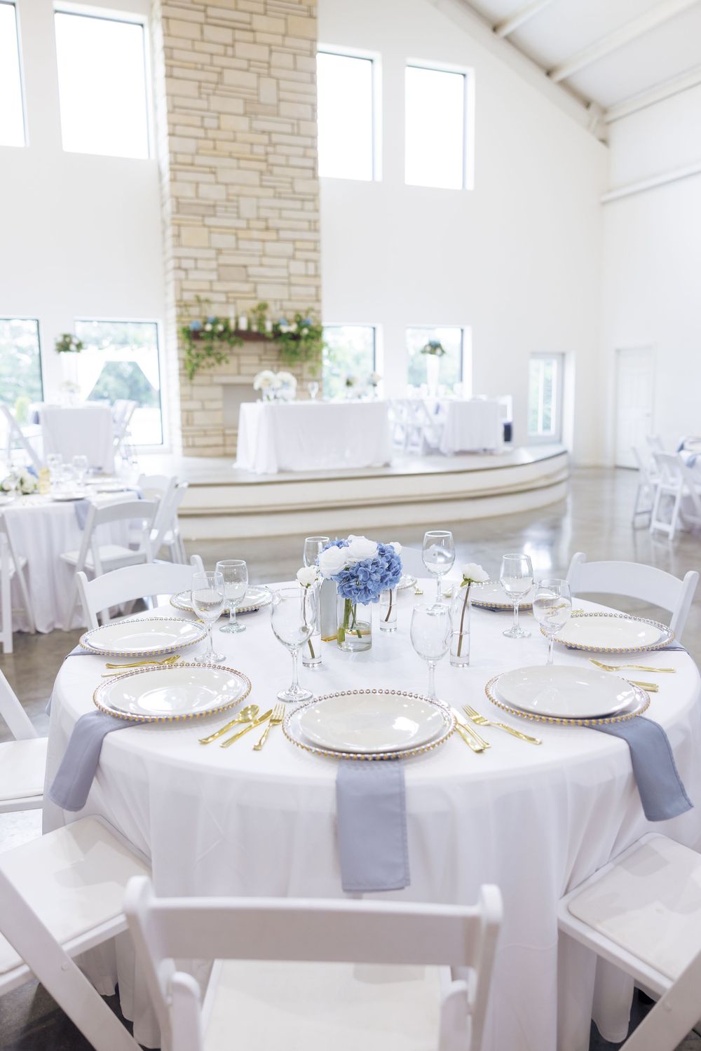 Elegant wedding reception hall with white round tables, gold chargers, blue hydrangea centerpieces, and white folding chairs.
