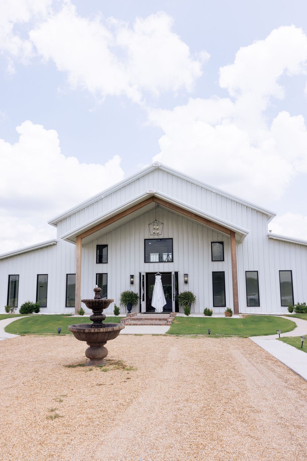 White barn wedding venue with a fountain, gabled roof, and a bridal gown hanging at the entrance under a cloudy sky.