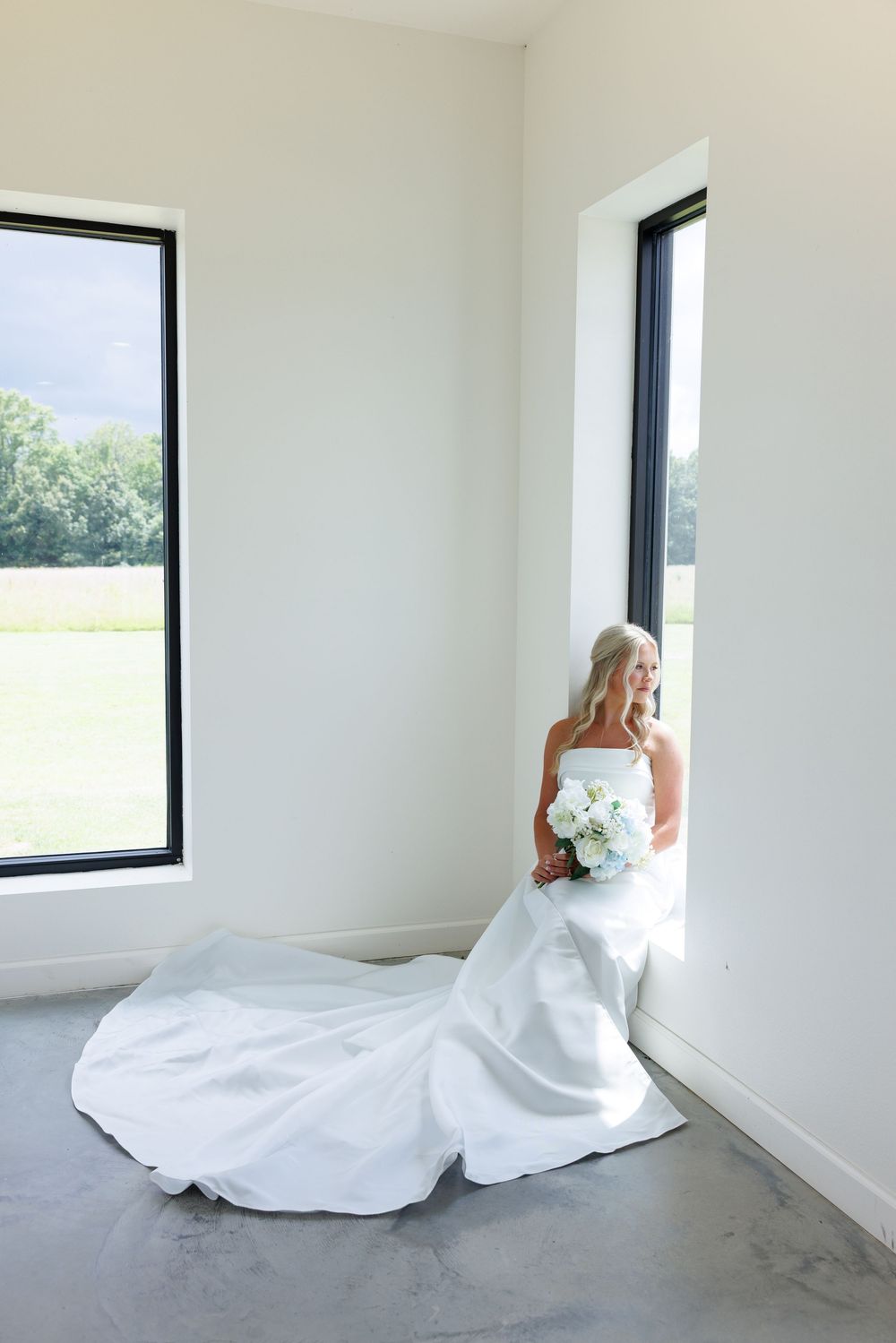 Bride in white strapless gown with long train holds bouquet by tall windows in bright modern venue.