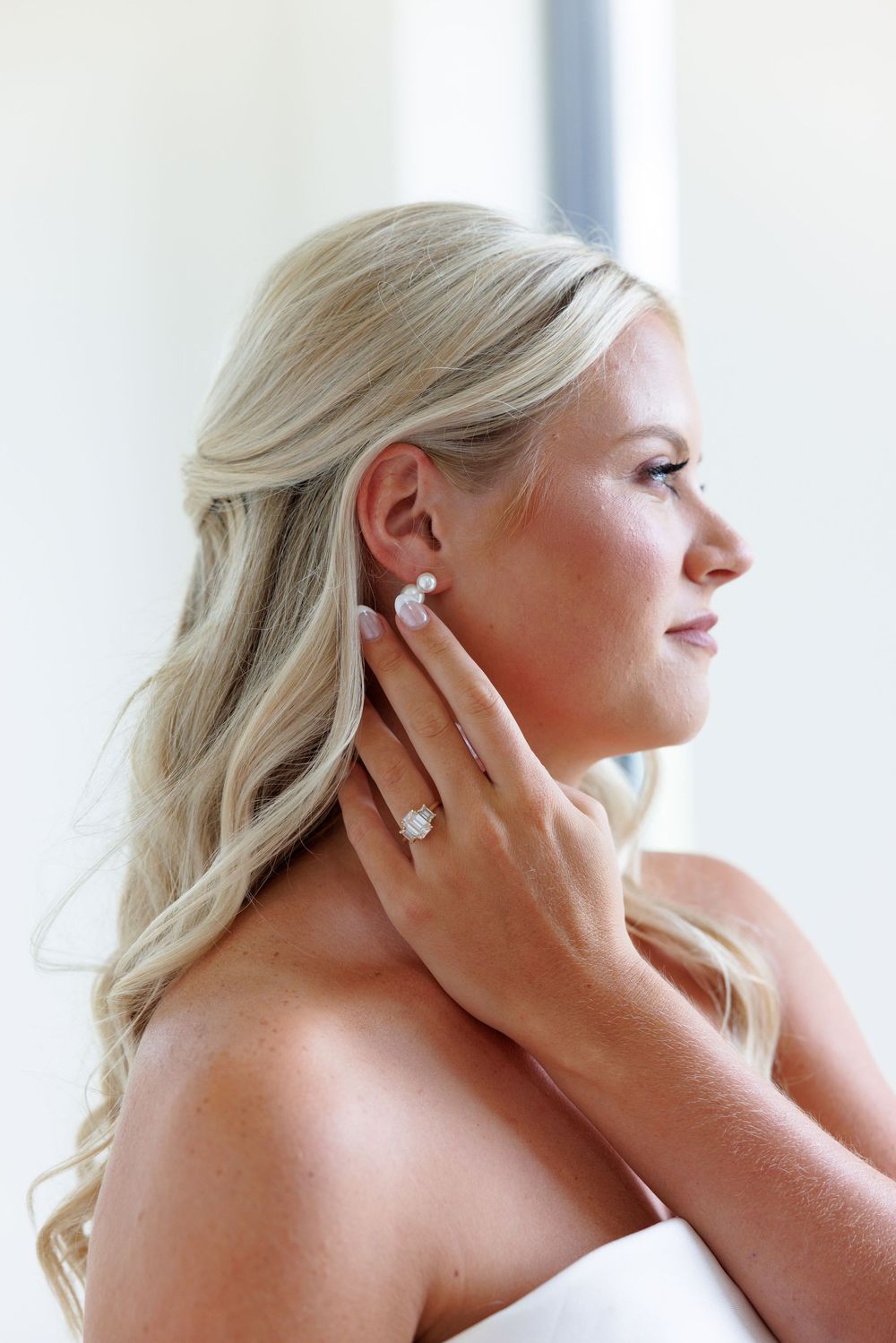 Blonde bride adjusting pearl earring, showcasing elegant diamond ring, soft waves, and strapless gown in bridal portrait.