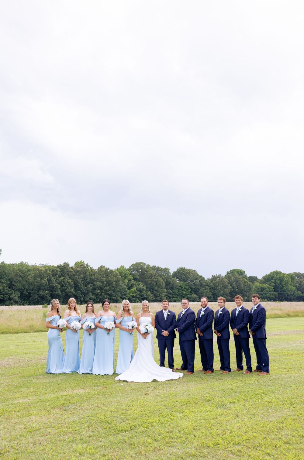 Wedding party posing outdoors with bridesmaids in light blue gowns and groomsmen in navy suits on a green field.
