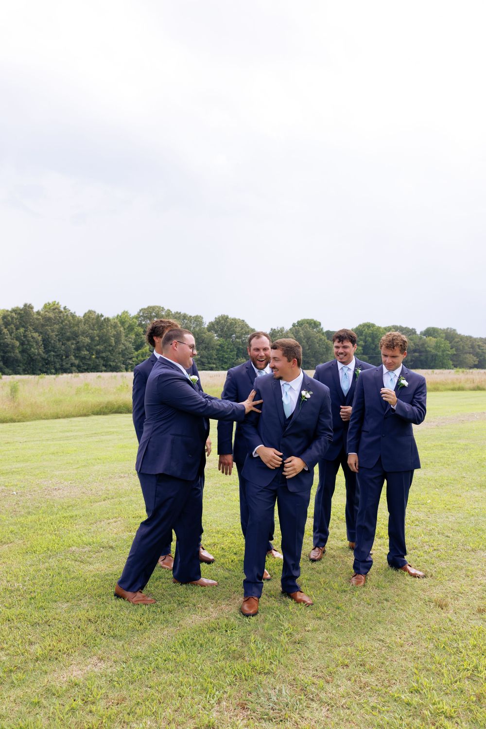Groom and groomsmen in navy suits laughing together on a green field at an outdoor wedding celebration.