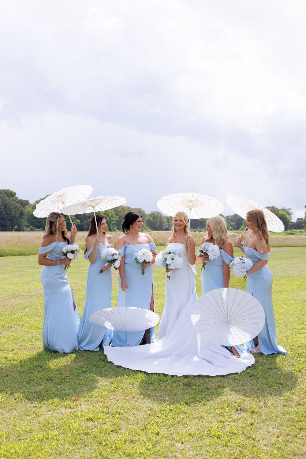 Bridesmaids in light blue gowns holding white parasols and bouquets surround the bride on a green lawn.