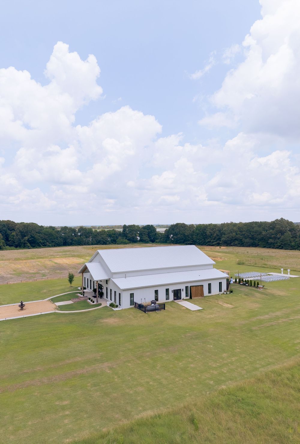 Aerial view of a white barn wedding venue surrounded by lush green fields and trees under a partly cloudy sky.