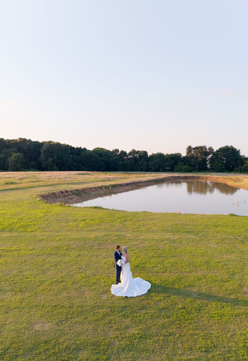 Aerial view of bride and groom embracing on lush green field beside a serene pond at golden hour.