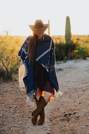 A figure in a blue poncho and cowboy boots stands against a desert landscape with saguaro cactus at sunset.