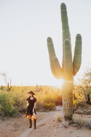 A silhouetted figure in western wear stands beside a towering saguaro cactus during golden hour in the desert.