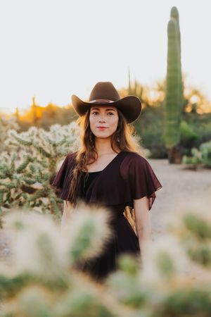 A western-styled portrait with black dress and cowboy hat against desert cacti and golden light.