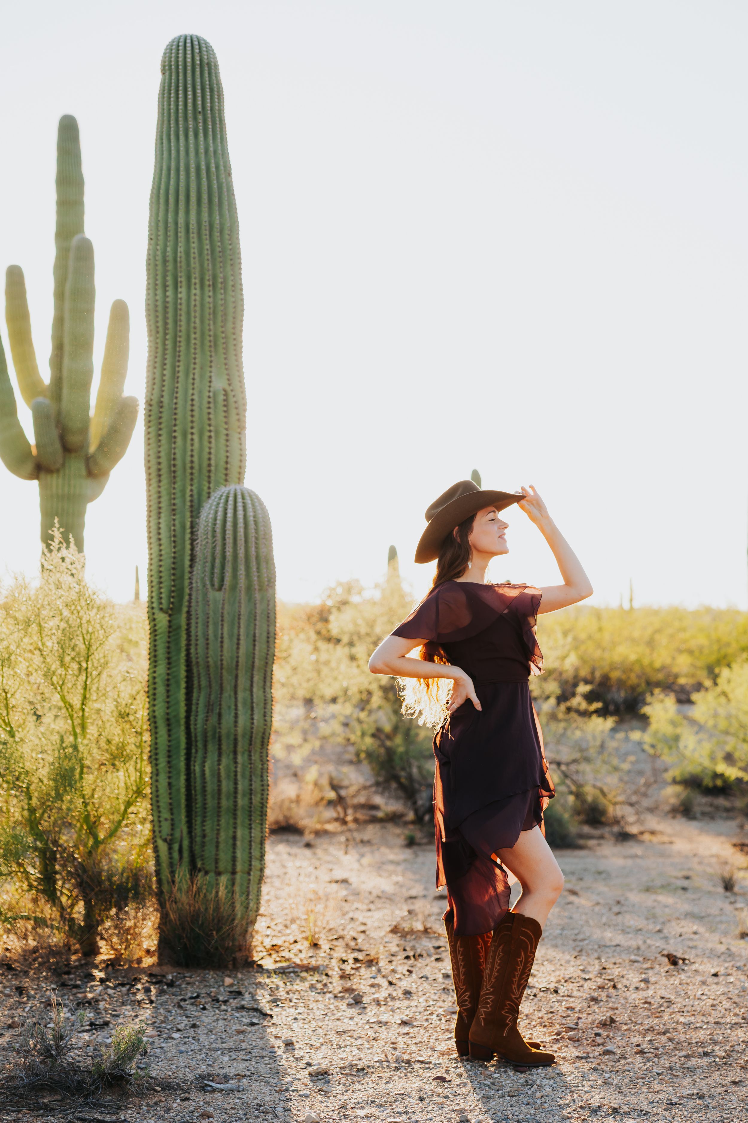 Cowgirl in brown boots poses playfully near saguaro cacti during desert photoshoot.