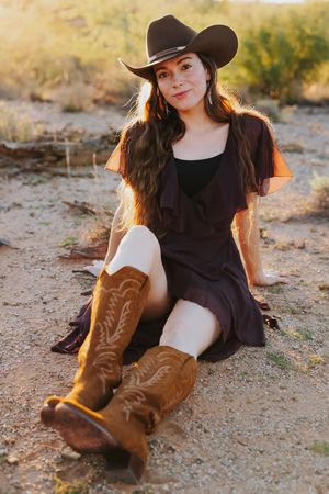 A rustic desert photoshoot showcasing brown cowboy boots and a black dress on sandy ground.