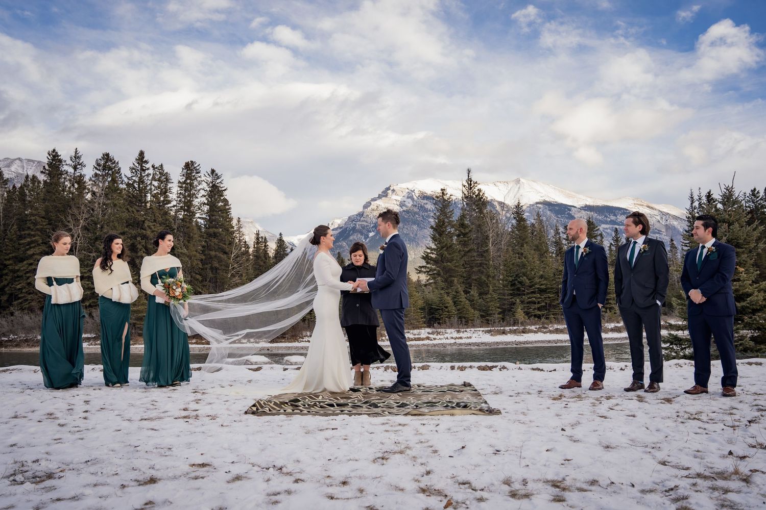 Wedding ceremony in winter mountain landscape with flowing veil and forest backdrop.