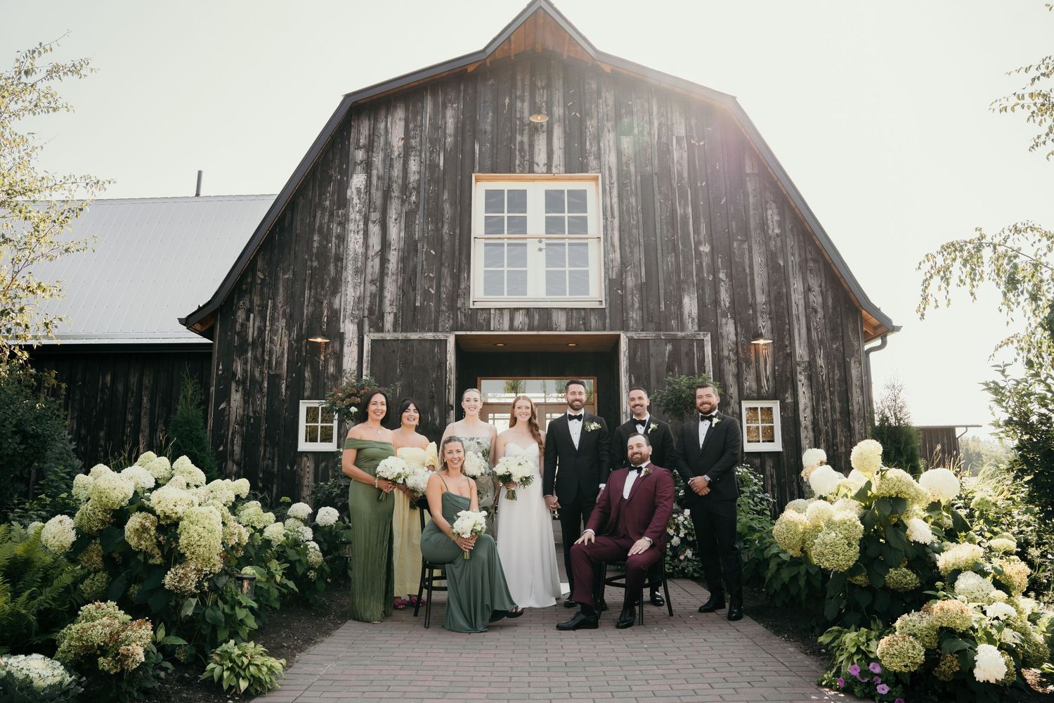 Wedding party poses in front of a rustic barn with white hydrangeas blooming in the foreground.