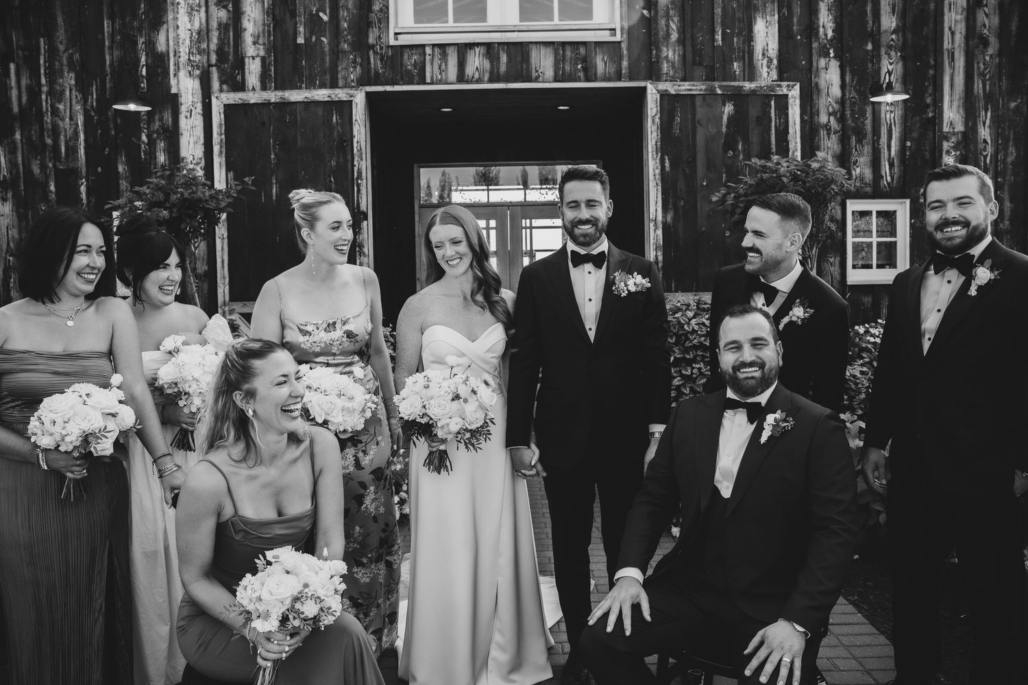 Black and white portrait of wedding party gathered in front of a rustic wooden barn entrance.