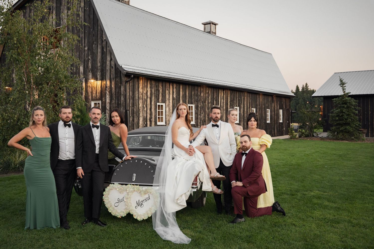 Wedding party in formal attire poses with vintage car outside rustic barn venue.