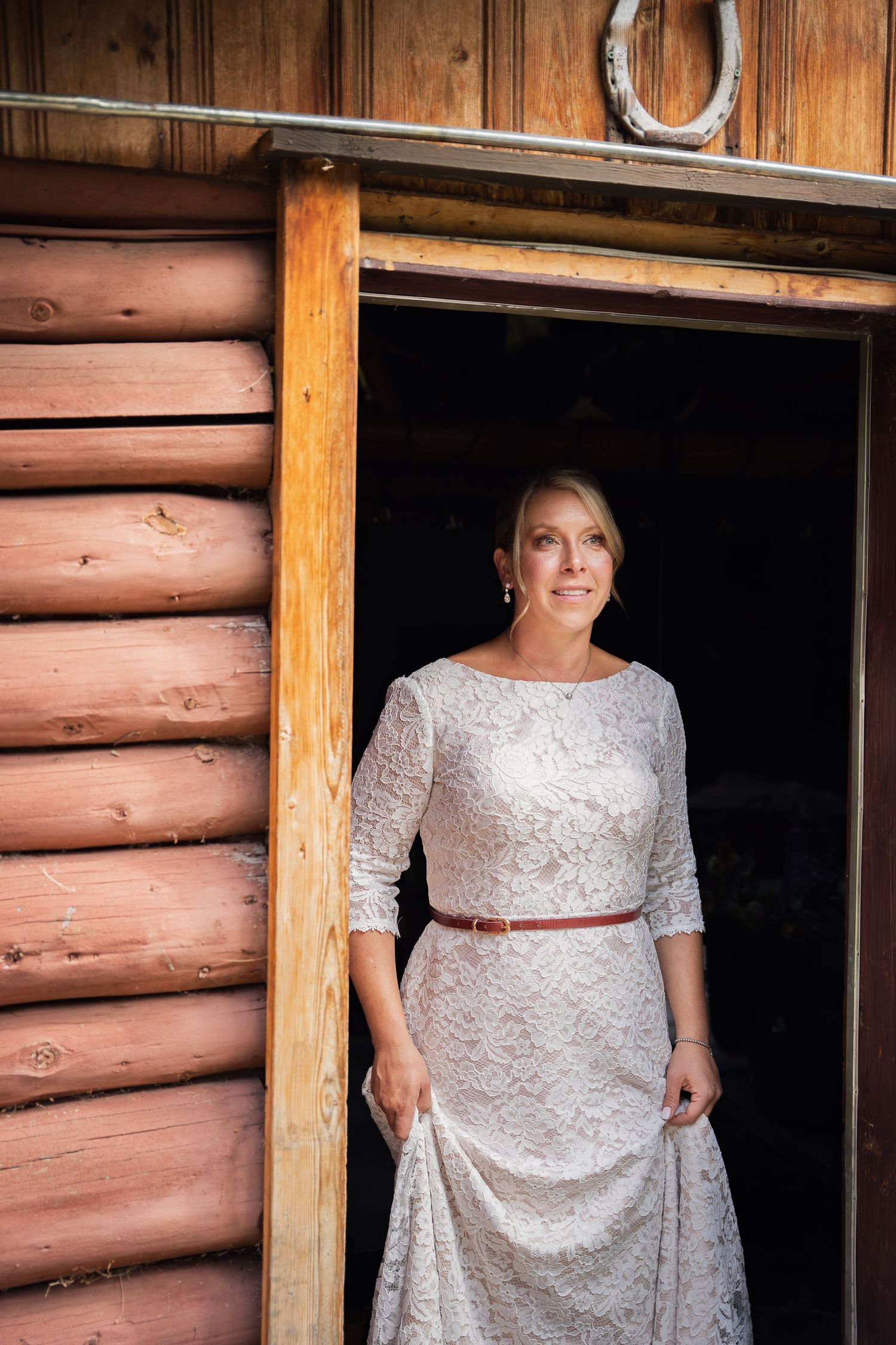 A white lace dress with a brown belt is worn in the doorway of a rustic log cabin.