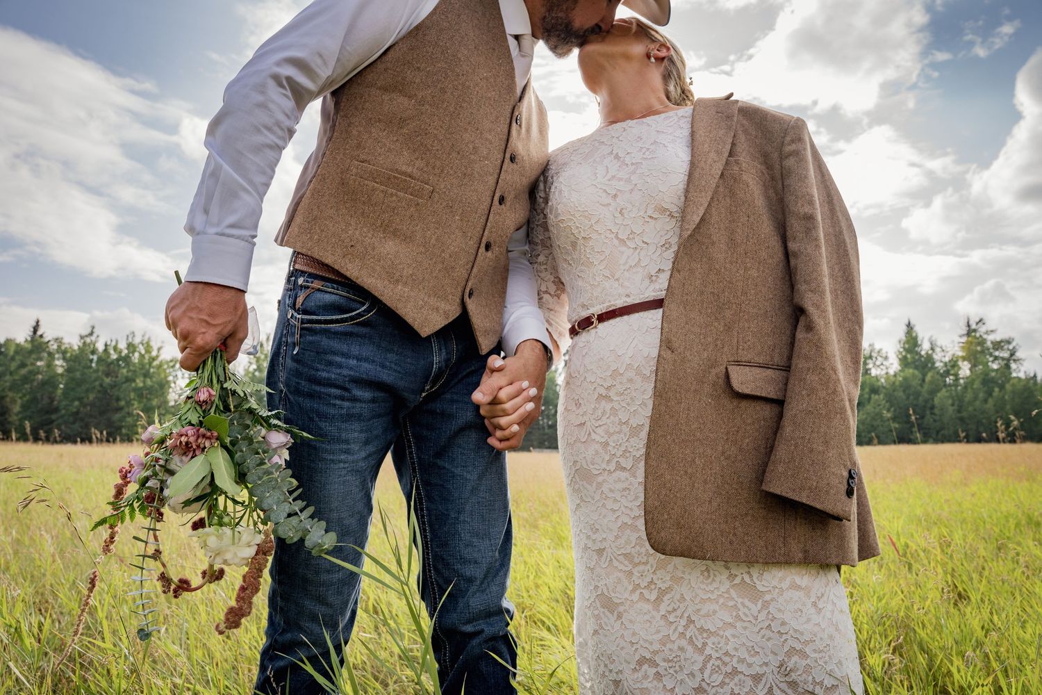 A couple holds hands in a sunny field while holding a brown tweed jacket and eucalyptus bouquet, country vibes, country wedding