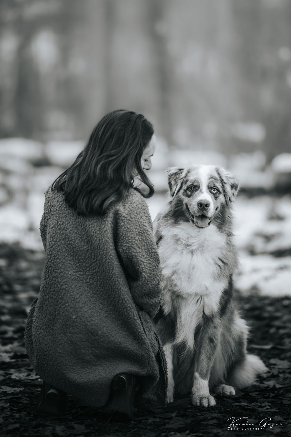 A woman in a dark coat stands in a snowy forest with a dog, likely a golden retriever, by her side.