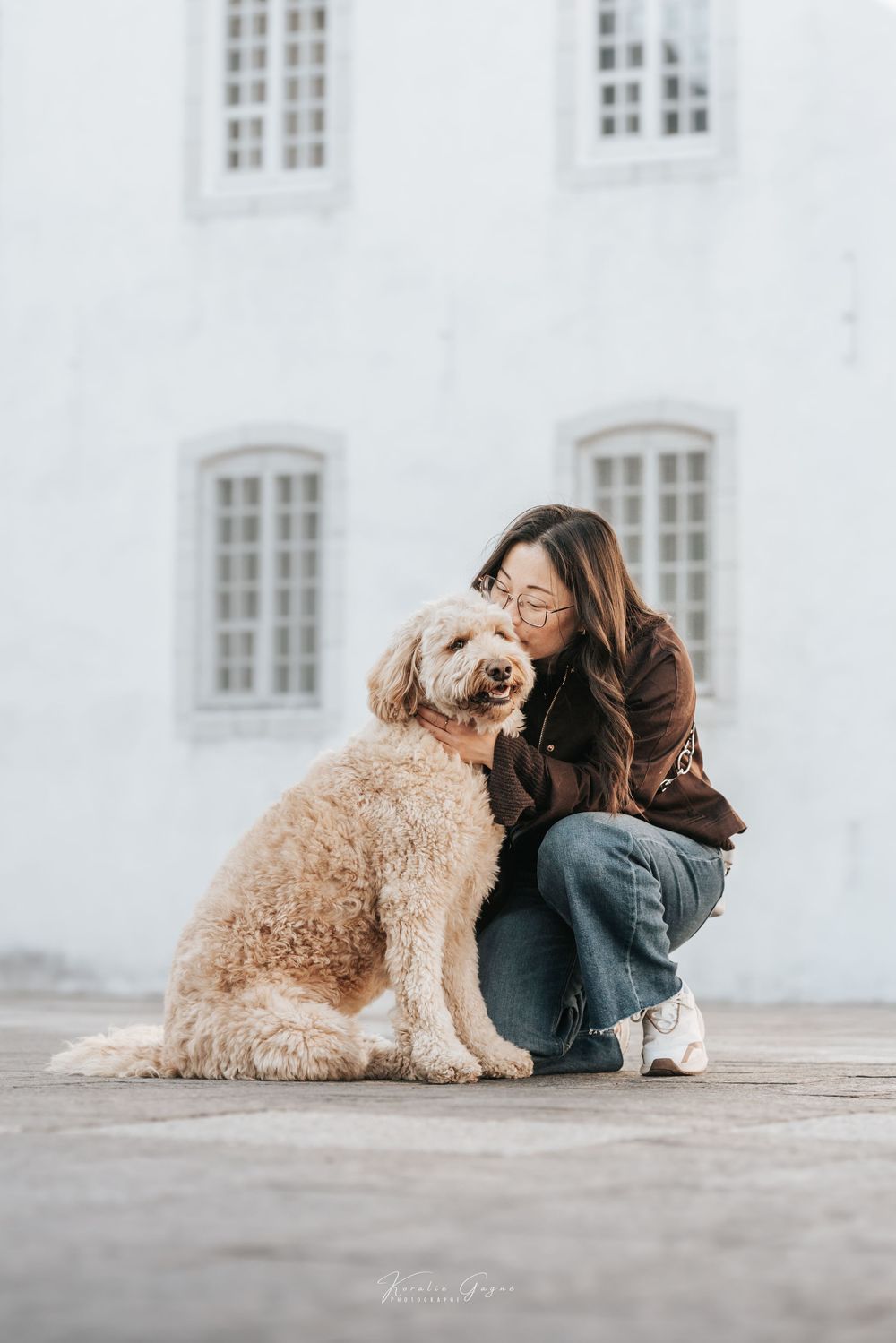 Woman in brown jacket and jeans crouching beside a fluffy golden doodle dog in front of a white building.