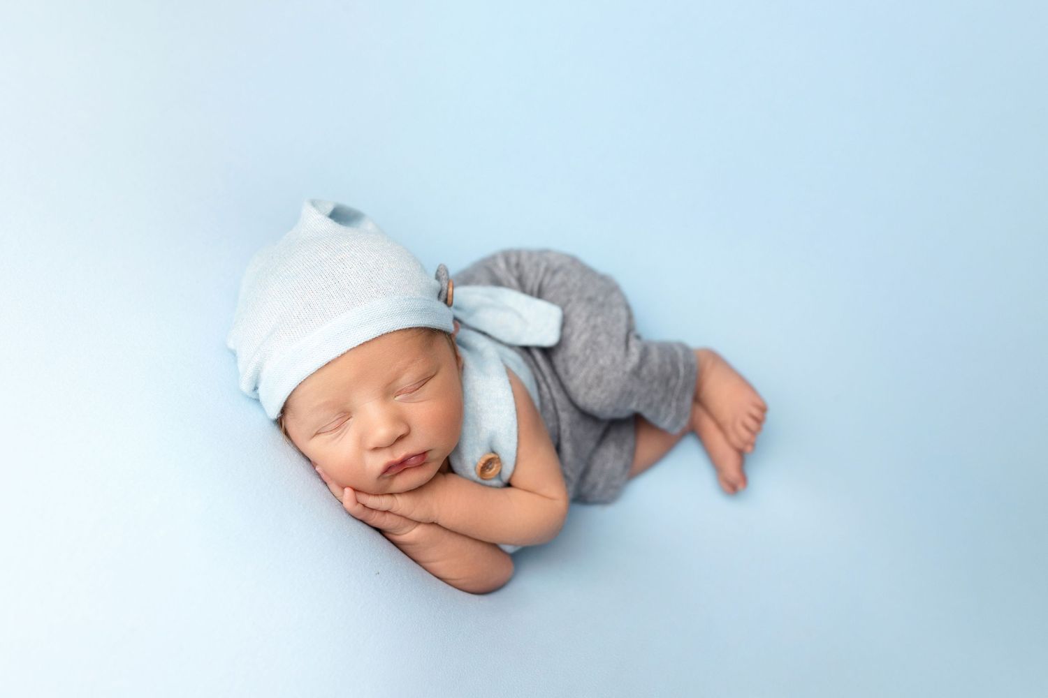 Sleeping newborn wearing blue and grey outfit with matching cap on light blue background.