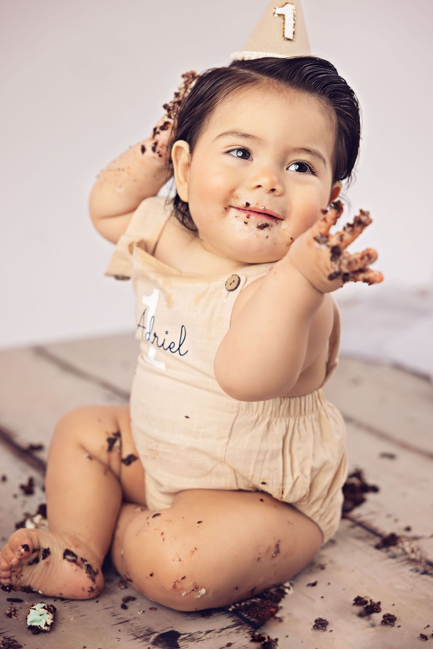Baby in beige romper playing with cake and frosting during first birthday celebration.