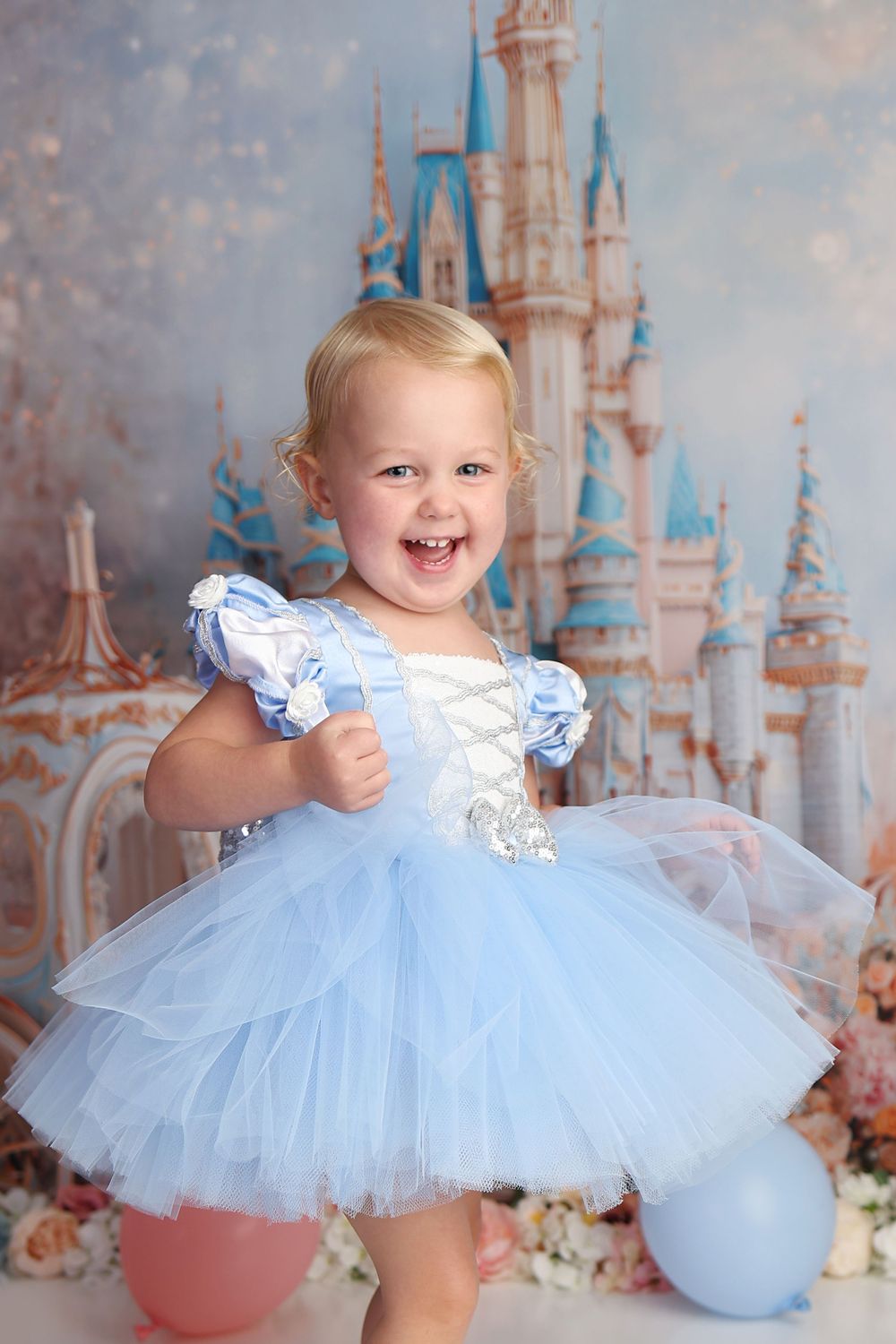 Smiling toddler girl in blue Cinderella tutu dress poses before a Disney castle backdrop with balloons.