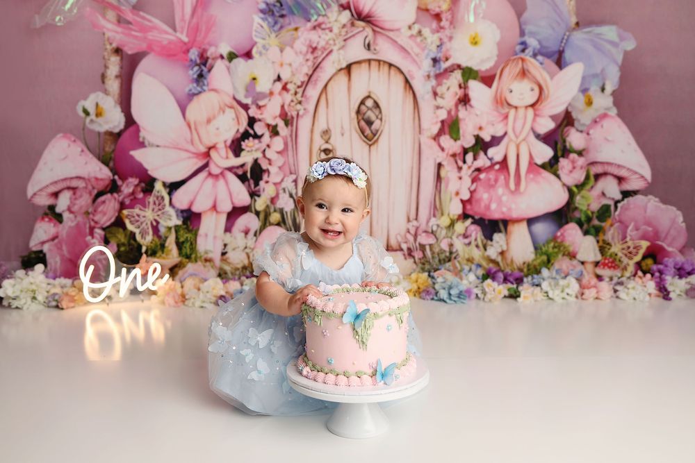 Baby girl in blue dress smiles at first birthday cake smash with fairy tale backdrop and glowing One sign.