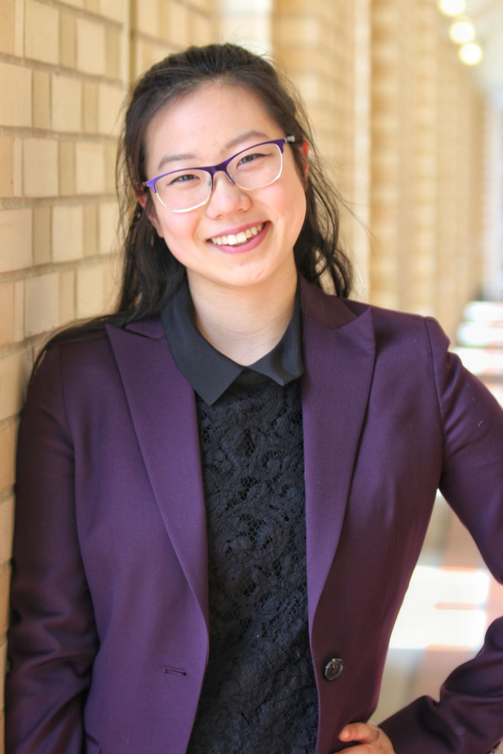 Professional wearing glasses and a purple blazer poses against a brick wall.