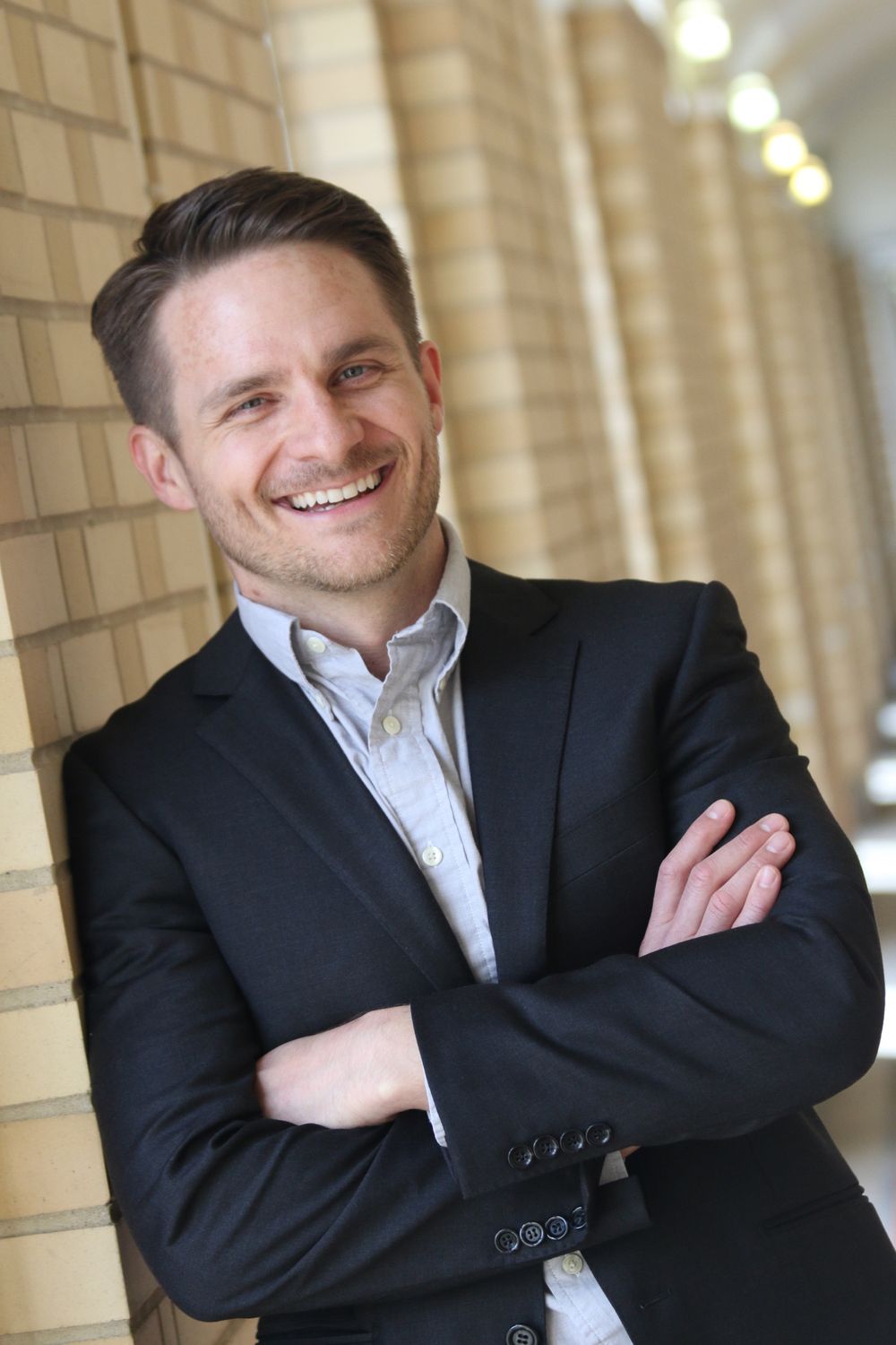 Professional portrait of a person in a dark suit and white shirt leaning against a brick wall.