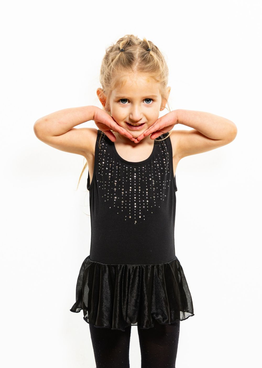 A young dancer in a black sparkly leotard with tulle skirt poses playfully with hands near face against white background.