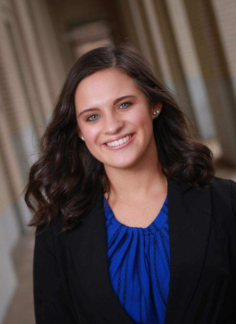 Professional headshot of a person in a blue blouse and black blazer with dark wavy hair smiling warmly.