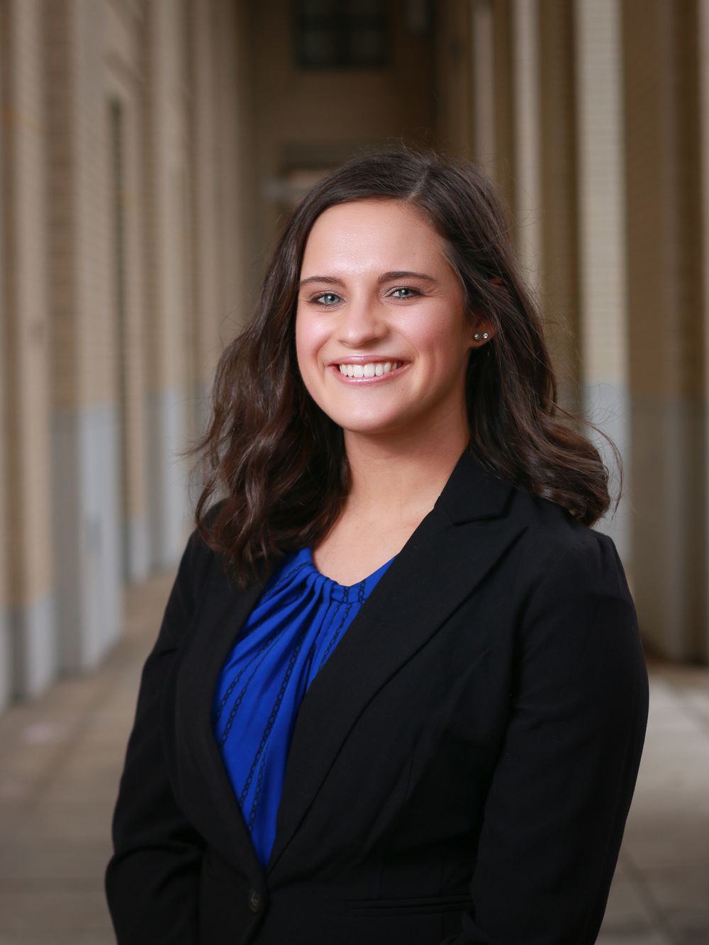 Professional headshot of a person in a black blazer and blue blouse against a columned background.