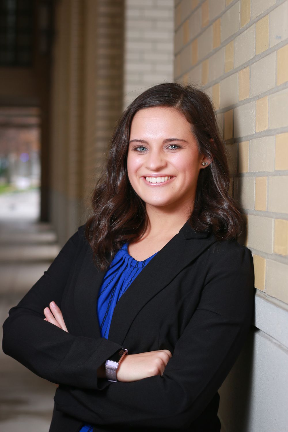Professional portrait of a person in business attire smiling with arms crossed against a brick wall.