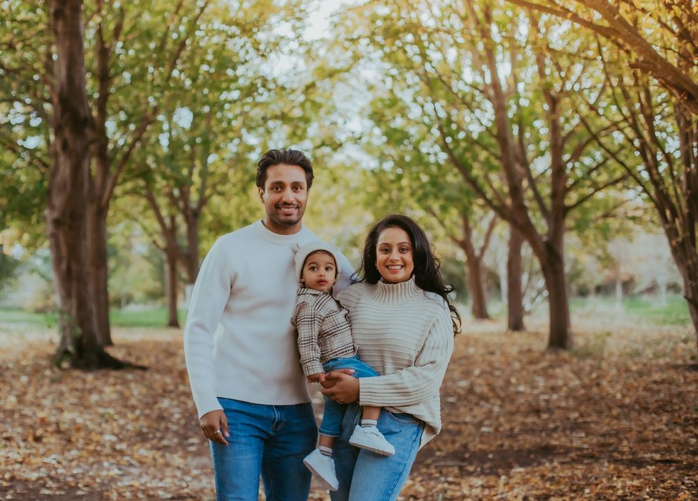 A happy family of three poses outdoors in a fall park, parents holding their baby among colorful autumn trees.